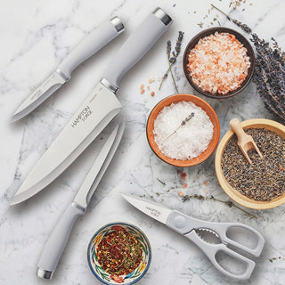 A set of taupe and grey kitchen utensils, including a knife, scissors, and a spatula, displayed with a natural pinewood cutlery block and a sprig of dill.